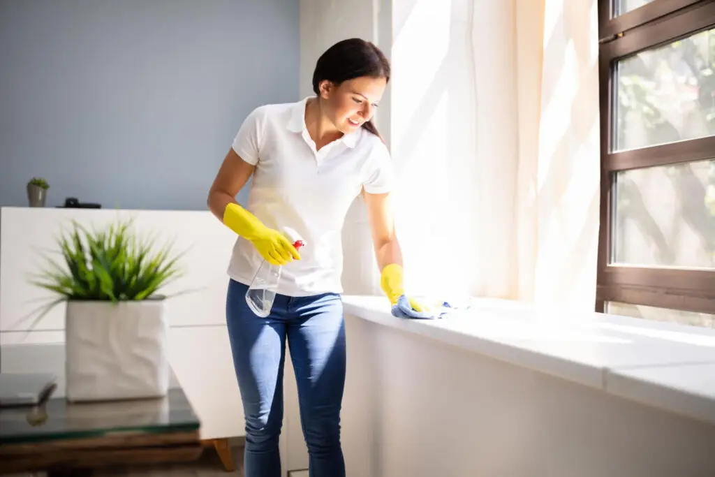 Woman wearing yellow gloves using a spray bottle and cloth to deep clean a white surface near a sunny window.