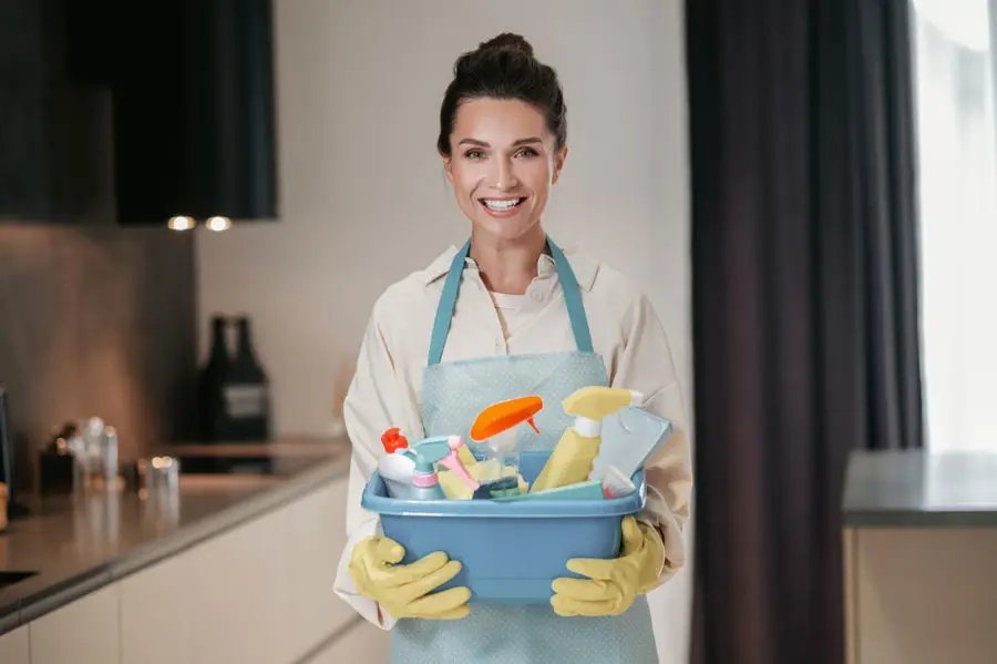 Smiling professional cleaner wearing an apron and gloves holding a bucket of supplies for a residential maid service.