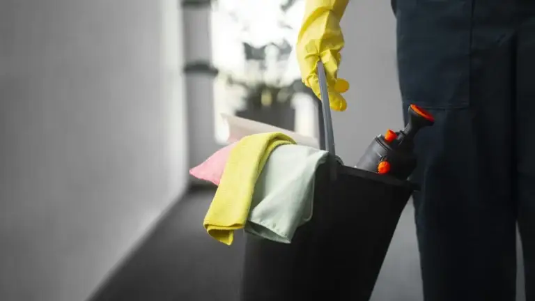 Cleaner in yellow gloves holding a bucket with microfiber cloths and spray bottle